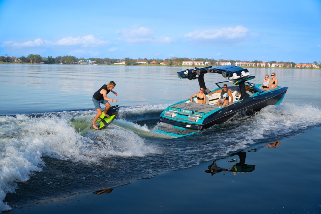 Person wakeboarding behind a turquoise motorboat as it speeds across a calm lake with riders watching from the boat
